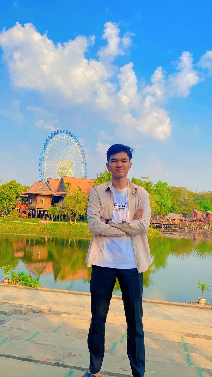 Kimlong KUN standing by a lake, with a Ferris wheel and wooden houses behind